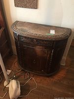 Full view of the vintage demilune wooden cabinet showing the carved top edge, drawer, door, and side details with visible scratches and wear.