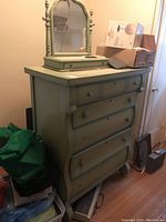 View of the green painted wood dresser with 7 drawers and mounted mirror, showing overall shape and condition in a corner of a room with boxes nearby.