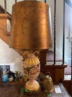 Table lamp with large sculpted ceramic base with gold-colored lampshade alongside small brass bell on wooden surface.