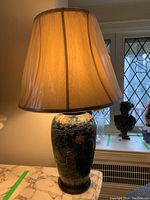 Table lamp illuminated showing ceramic base with dragon and floral pattern under beige fabric lamp shade in plastic wrap, placed on marble surface near window.