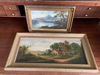 Two framed acrylic paintings showing a rural cottage scene and a mountain lake scene in horizontal golden frames with worn edges