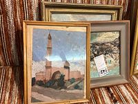 Three vintage framed paintings positioned together showing frames and parts of scenes, one with a small tower under a blue sky