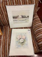 Two framed watercolours placed on a striped fabric chair, one depicting a barn scene and the other a flower bouquet, both signed Hans