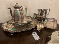 Set of silver plated serving items on a wooden surface, including coffee urn, gravy boat, two serving trays, double handle jug, and stainless steel jug.