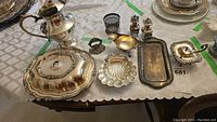 Overview photo showing all silver plated serving dishes including lidded dish, pitcher, salt and pepper shakers, small tray and other dishes arranged on a table with white cloth.