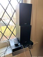 Two black Sony speakers stacked on top of Sony receiver on a window sill, with remote control resting on top of receiver.