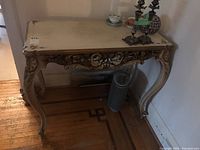 Full side view of the ornate vintage table showing cream-colored tabletop with floral decorations and curved legs with carvings.