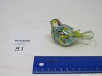 Glass bird paperweight with colorful internal patterns shown from above beside a blue ruler indicating approximate size of 8 cm in length.