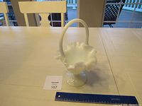 White glass basket with handle and ruffled edge on pedestal base on a wooden table with a ruler for scale, showing size and condition.