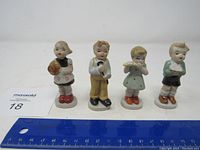 Four porcelain child figurines arranged in a row on a white surface with a blue ruler in front for scale. Includes a girl holding a hat, boy with a cane, girl playing a flute, and boy with arms crossed.