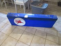 Blue rectangular Mobil sign with large red Pegasus logo on a white circular background placed on tile floor, showing overall appearance and mounting chains on top.