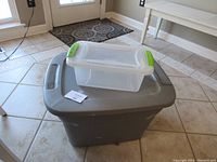 Large gray plastic storage container with lid and smaller clear container with green handles on top, placed on tiled floor near door
