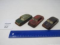 Three old tin toy cars displayed with a blue ruler to indicate size. Cars vary in color: green with a checkered roof, red, dark blue with white stripes. Signs of rust and wear are visible.