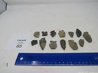 Top-down view of all 13 stone arrowheads laid out in a single row with ruler for scale.