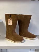 Side view of two mid-calf brown suede Bearpaw boots with white rubber soles and tags attached, showing the exterior and shape of boots.