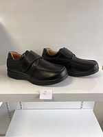 Side-angle view of men's black comfort shoes with hook and loop closure strap on a white shelf.
