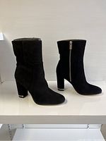 Side view of the pair of black suede ankle booties with visible silver zipper and block heels on white shelf.