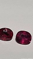 Close-up photo showing two oval-shaped hot pink-red sapphires with visible facets and some inclusions.