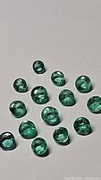 Top view of 16 small round calibrated emerald gemstones scattered on a white surface showing their green color and faceted cut.