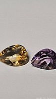 Pair of pear-shaped faceted gemstones, citrine (yellow) and amethyst (purple), shown on white background.