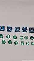 Photo showing five square-cut London blue topaz and multiple round-cut emerald gemstones loose on a flat surface.
