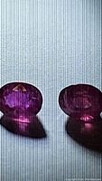 Two oval cut hot pink sapphire gemstones on a white lined surface, showing facets and reflections with visible inclusions.