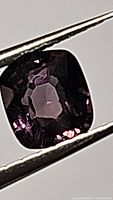 Close-up view of 1.36 carat deep purple cushion-cut spinel held by tweezers, showing facets and clarity.