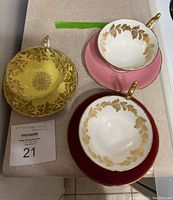 Top-down view of three tea cups and saucers in yellow, white with pink saucer, and white with maroon saucer, showing gold leaf and floral patterns.