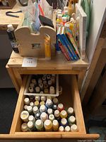 Photo of wooden caddy holding brushes, paint bottles, canvas, pastel sets, and poly bags on a wooden table with a drawer holding additional paint bottles.