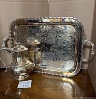 Silver plated coffee pot next to a large ornate serving tray on a wooden surface