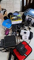 Lot of electronics on a table showing various devices including Garmin GPS, Sony CD player, radio, alarm clock, earbuds, walkie talkies and accessories.