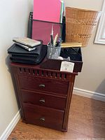 Photo showing the red wooden filing cabinet with three drawers, metal handles, and various office supplies including pens, notebooks, a small wooden brush, and organizing cases placed on top of the cabinet
