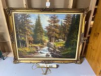 Front view of the framed landscape painting showing ornate gold frame, forest scene with running stream and rocks in detail.