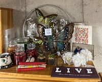 Display of three large metal butterfly wall art pieces mounted on metal rings, surrounded by assorted scented candles in glass jars, two small artificial succulent plants, white fabric flowers, and decorative signs including a 'LIVE' plaque and printed papers.