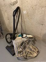 Tristar vacuum with hose, plastic bag with felt hangers, metal closet organizer pieces on basement floor.