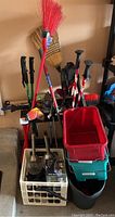 Photo showing several hiking poles, brooms including a red angled broom, a red plastic bucket in a black wheeled garden cart, and house cleaning tools such as dustpan, scrapers, and dusters.