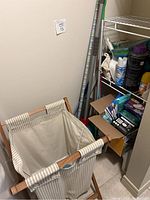 Photo showing laundry basket with striped fabric and wooden frame, mop and broom handles next to it, metal shelf with various cleaning products.