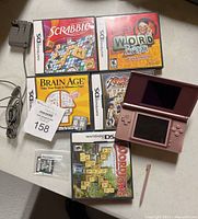 Pink Nintendo DS Lite console, stylus, six Nintendo DS games and charging cord, laid out on a surface, well lit