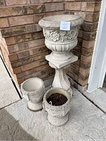 Full view of the three ceramic planters and the pedestal arranged on stone surface near a brick wall.