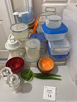 Wide view of a large assortment of plastic kitchenware items including bowls, plates, storage containers, a shaker bottle, and salt and pepper shakers arranged on a table.