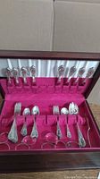 Wooden flatware chest open to reveal red velvet interior holding ornate silver-plated forks and serving spoons