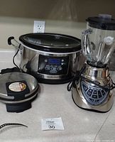 Three small kitchen appliances on countertop: Hamilton Beach slow cooker, Oster blender, and Cuisinart waffle maker.