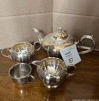 Photo of the complete four-piece tea set featuring the teapot, creamer, sugar bowl, and a small metal cup on a wooden table.
