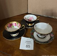 Photo showing all three teacups and saucers on a wooden table with a cardboard backdrop and a MaxSold lot number tag.