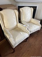 Photo showing two matching wingback armchairs side by side on a wooden floor with a fireplace in the background.