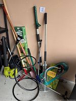 Collection of various yard tools leaning against a wall including sprinkler, shovels, rake, broom, and hedger tools