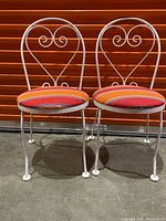 Two white metal framed bistro chairs with red-orange striped cushioned seats in front of an orange roll-up door. Showing full front view of both chairs with decorative scroll heart-shaped backrests.