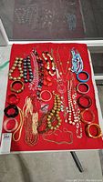 Overview of costume jewelry on red cloth including numerous necklaces, bangles, and bracelets of various colors and materials.