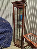 Tall dark wood display cabinet with a glass door and four glass adjustable shelves, standing against a corrugated metal background.