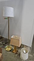 Wide view showing brass floor lamp with white cutout patterned lampshade, acrylic table lamp with rectangular beige lampshade, plus extra lampshades and lamp harps.
